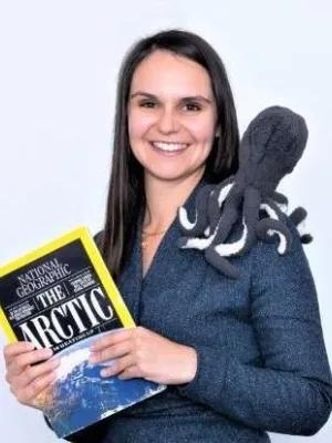 A woman in a blue shirt smiling in front og a white background. She is holding a magazine and a toy octopus