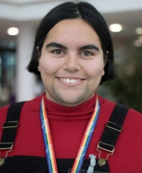 Cara is smiling at the camera wearing a red top and brown dungarees, with a pride flag lanyard around her neck.