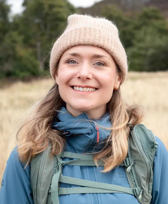 A woman smiles at the camera. She is wearing a warm hat and coat and standing in front of a field