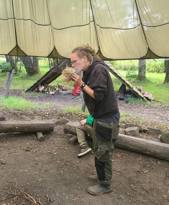 A woman under a large fabric shelter blows into some straw in her hands. 