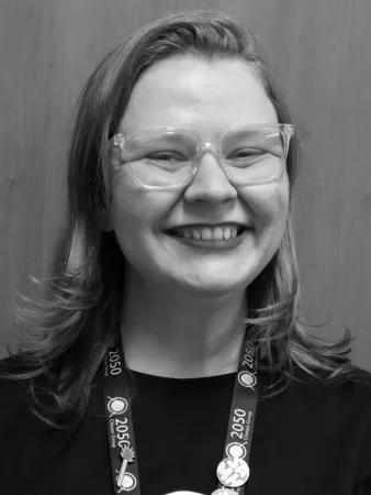 A black and white photo. A woman in glasses with shoulder length hair and a black t-shirt smiles at the camera