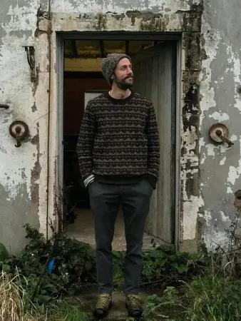 A man stands in front of a building with white paint cracking. He is wearing a knitted sweater and hat and looking to the right