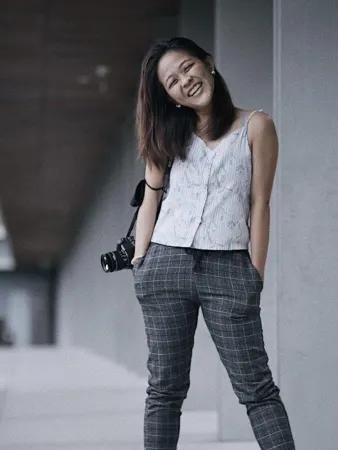 A woman stands in front of a grey building. She has long, dark hair and a camera over her right shoulder