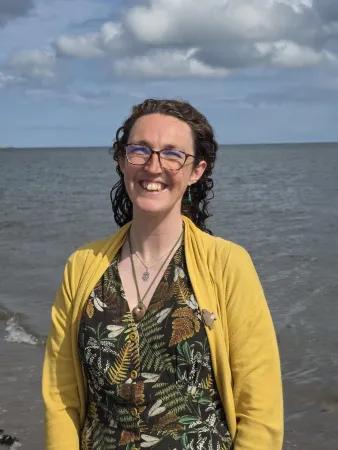Lucy standing in front of the sea. She has glasses, long curly hair and is wearing a yellow cardigan