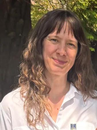 A woman with long brown hair and a white t-shirt stands in front of a tree smiling at the camera