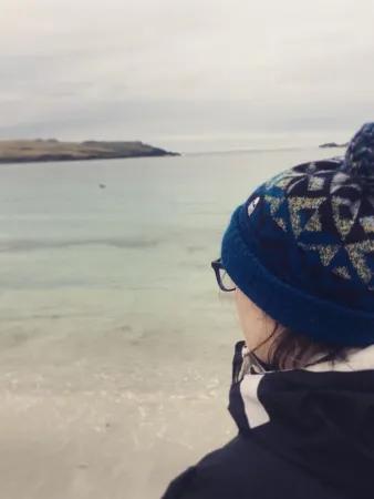 A woman looks out onto a calm sea. We see the back of her head wearing a blue bobble hat and black coat