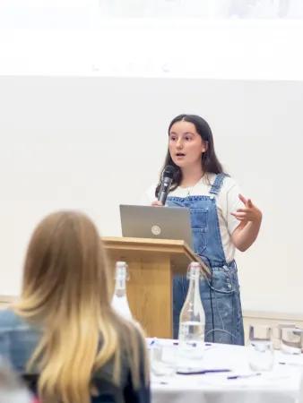 A woman with long dark hair and in blue dungarees is at a lectern speaking to a number of seated people with their backs to the camera
