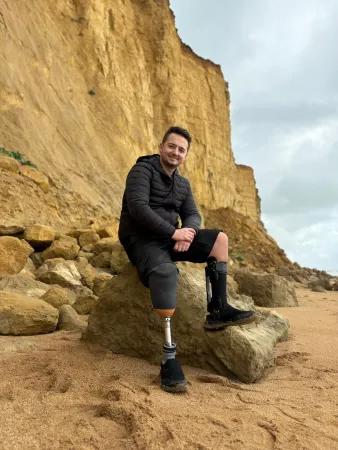 A man with short hair and a black coat sits on a rock on a beach with high cliffs behind him. He has a prosthetic right leg and a brace on his left leg